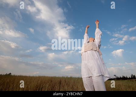 Young woman wearing white dress with hands up in the sky. Caucasian woman dancing outdoors in nature Stock Photo