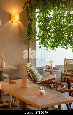 Cozy café corner with a rattan chair, a small round table featuring coffee, pastries, and a vase with dried flowers, surrounded by lush hanging plants Stock Photo