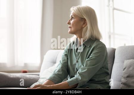 Pensive sad senior female sitting on couch and looking away Stock Photo