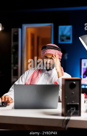 Tired male entrepreneur in Arabic attire browses the internet on a laptop, frustrated, he touches his forehead. Despite feeling sick, Middle Eastern man remains committed to his work in modern office. Stock Photo