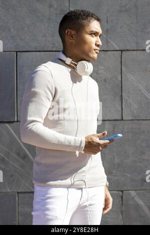 Vertical portrait of serious black latin man in white dress looking to the side with headphones and cell phone, vertical wall background Stock Photo
