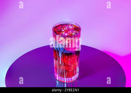 Tall glass of cold beverage with ice cubes against vibrant pink and purple light on black reflective surface. Stock Photo