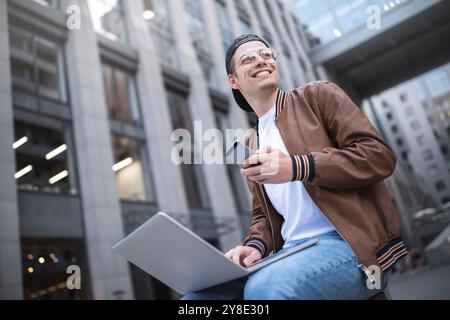 Happy handsome student looking at computer screen while typing something on laptop keyboard during work break outdoors, teenager looking at the notebo Stock Photo