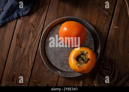 Two ripe persimmons on a round metal plate, with a rustic wooden background and dried leaves. Stock Photo