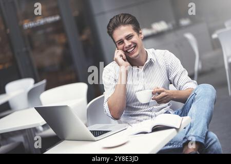 Handsome caucasian young man with phone working on laptop and smiling while enjoying coffee in cafe Stock Photo