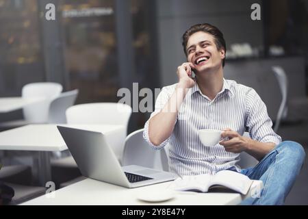 Handsome caucasian young man with phone working on laptop and smiling while enjoying coffee in cafe Stock Photo