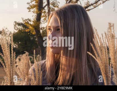 A woman with long, blonde hair stands in a field of tall grass, looking off to the side Stock Photo