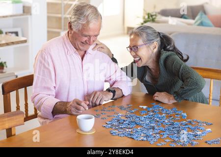 Happy senior diverse couple sitting at table and doing puzzles Stock Photo