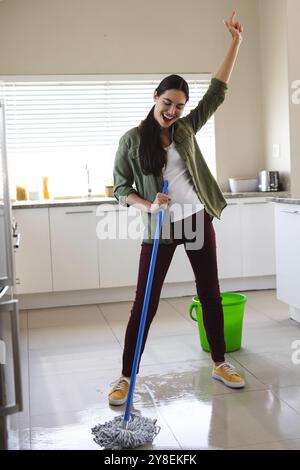 Cheerful caucasian young woman with hand raised dancing and cleaning floor with wet mop at home Stock Photo