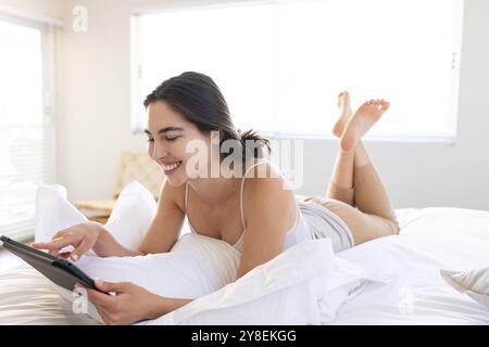 Caucasian smiling young woman using digital tablet while lying on bed at home, copy space Stock Photo