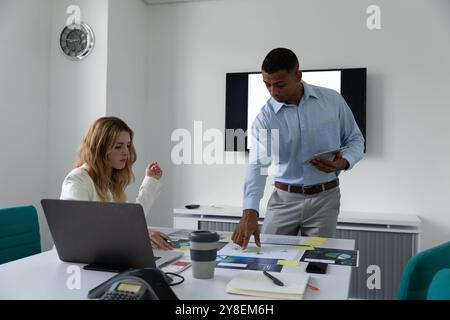 Front view of a young African American man standing holding a tablet computer and talking with a young Caucasian woman sitting at a desk, they are usi Stock Photo