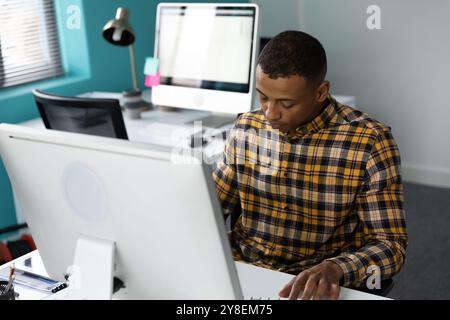 Front view of a young African American man sitting at a desk using a computer in the modern office of a creative business, with an empty workstation i Stock Photo