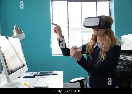 Side view of a young Cauasian woman sitting at a desk wearing a VR headset and with one hand raised and finger pointing in the modern office of a crea Stock Photo