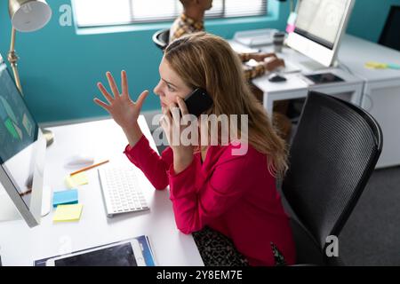 Side view close up a young Caucasian woman sitting at a desk in front of a computer talking on a smartphone and gesturing with her hand raised, in the Stock Photo