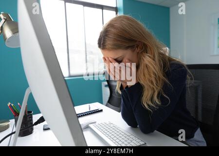 Side view of a young Caucasian woman sitting at a desk using a computer with her head in her hands in the modern office of a creative business Stock Photo