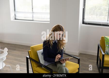 Side view of a young Caucasian woman sitting in an armchair using a tablet computer in the lounge area of a creative business Stock Photo