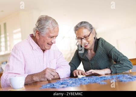 Happy senior diverse couple sitting at table and doing puzzles Stock Photo
