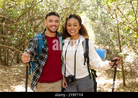 Portrait of happy biracial couple wearing backpacks and smiling in forest Stock Photo