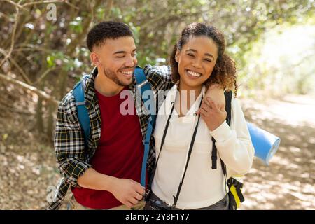 Portrait of happy biracial couple wearing backpacks and smiling in forest Stock Photo