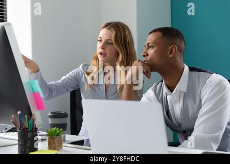 Side view close up of a young African American man using a laptop computer listening to a young Caucasian woman talking and pointing to a computer mon Stock Photo