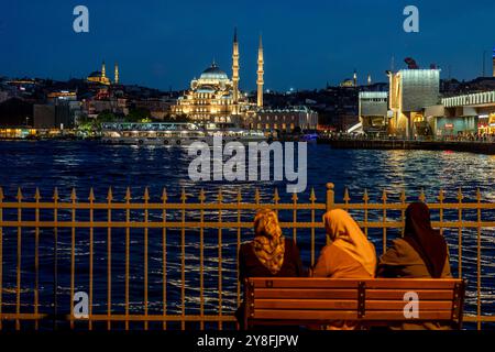 Istanbul. Turkiye. Three Muslim women sitting on a bench in front of the Bosphorus at dusk with the Yeni Cami Mosque on background Stock Photo