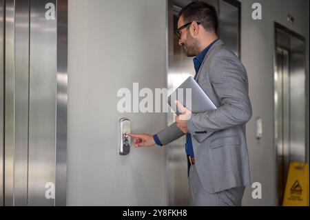 Side view of young businessman holding laptop and pushing button while waiting for elevator in office Stock Photo