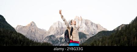 A woman stands triumphantly with her arms raised towards the sky, surrounded by towering mountain peaks and lush greenery. Dressed in a light jacket a Stock Photo
