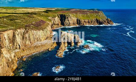 Aerial view of rugged cliffs meeting the ocean, with waves crashing against the rocks. The landscape features green hills and a clear blue sky, showca Stock Photo