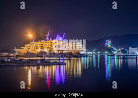 Night corniche promenade with illuminated Mutrah castle on the hill and highlighted sea bay with cruise ship and motor boats in the port, Muscat, sult Stock Photo