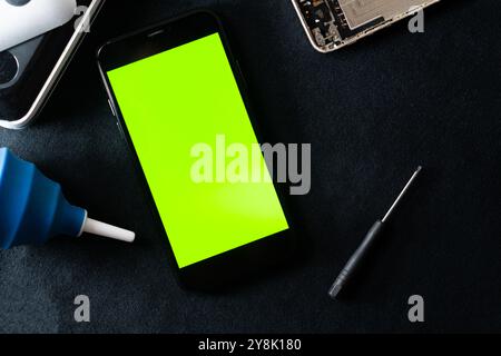 A smart phone with a bright green screen placed on a dark surface. Nearby, there is a blue glue bottle and a small screwdriver, suggesting a repair or Stock Photo