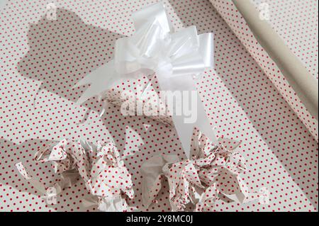Close-up of a gift wrapped in red polka dot paper with a white bow, surrounded by crumpled paper and wrapping roll, symbolizing holiday preparation an Stock Photo
