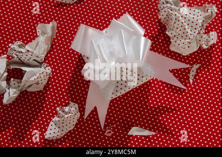 Gift box with white bow surrounded by crumpled wrapping paper on a red polka dot background, symbolizing celebration and surprises. Stock Photo