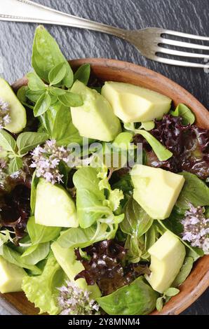 Top view at clay dish with salad of avocado, green and violet lettuce, lamb's lettuce and oregano flowers on slate stone tray with fork aside Stock Photo