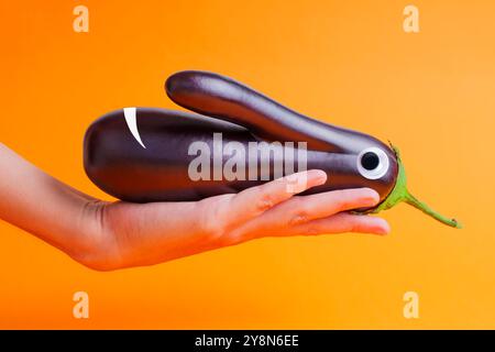 Hand presenting a playful eggplant character with googly eyes against a vibrant orange background. Cheerful and fun depiction perfect for food-related Stock Photo