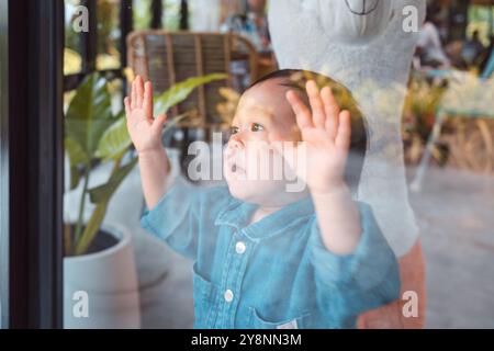 Cheerful asian toddler boy standing and clinging to the window glass at restaurant Stock Photo