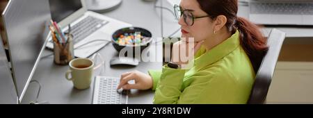Website header shot of young red haired female software engineer wearing green shirt and glasses focused on reading computer screen, while sitting at working desk in modern programmer office Stock Photo