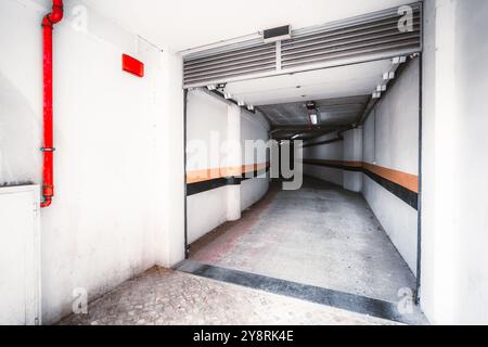A white underground garage entrance with curved walls, featuring orange and black horizontal lines, overhead lighting, a red fire pipe, and a paved st Stock Photo