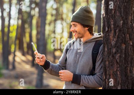 Positive guy leaning on tree in forest, using mobile phone Stock Photo