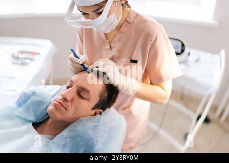 Top view of balding male patient with hair loss problem lying on couch, nurse marking with pencil new hairline on head before hair transplantation Stock Photo