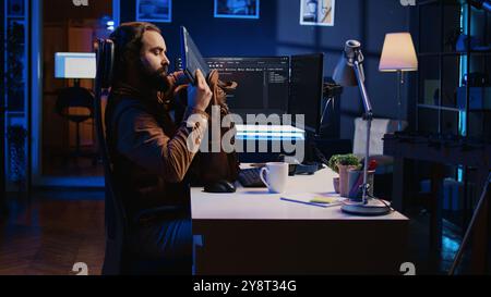 Software designer finishing remote job shift, packing backpack, leaving office after finishing coding project. IT professional putting laptop in bag, sitting up from desk chair, camera B Stock Photo