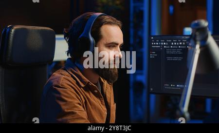 Smiling developer listening music while doing programming during remote job shift in home office. Upbeat IT admin enjoying audio podcast, working on PC in apartment late at night, close up Stock Photo