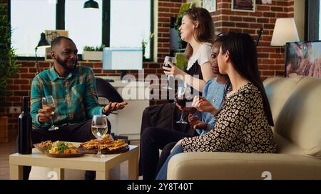 Culturally diverse friends talking, drinking alcoholic beverages and eating cheese, bread sticks from charcuterie board at home. Multicultural colleagues enjoying social life during weekend Stock Photo