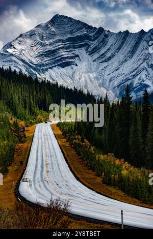 Autumn road through Kananaskis: winding path, golden trees, towering mountains, cloudy skies. Stock Photo