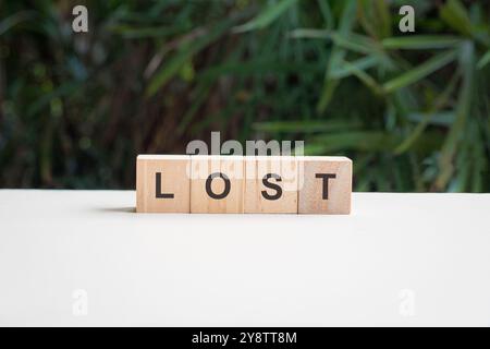 Lost, word written on square wood block, on top of white table with palm plant at the background. Stock Photo