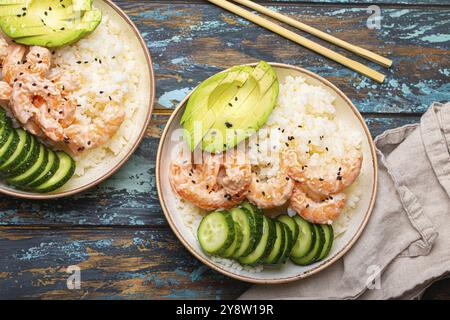 Two white ceramic bowls with rice, shrimps, avocado, vegetables and sesame seeds and chopsticks on colourful rustic wooden background top view. Health Stock Photo