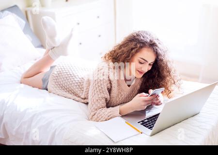 University student attending morning online class on laptop, lying on bed in dorm room. Stock Photo