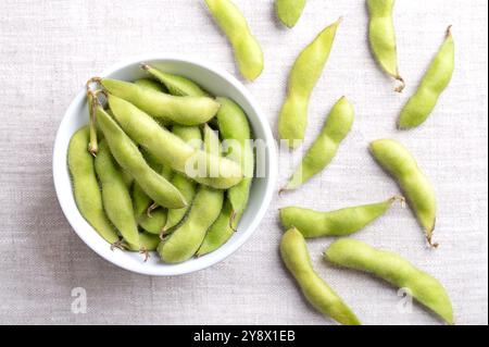 Fresh edamame, green soybeans in the pod, in a white bowl, on linen. Raw, immature soybeans, Glycine max, used boiled or steamed and salted. Stock Photo