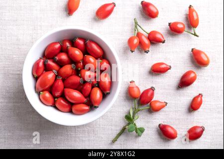 Fresh rose hips in a white bowl on linen. Also rose haw or rose hep, intense red ripe fruits, used for herbal teas, jam and can be eaten raw. Stock Photo