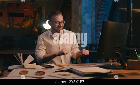 Caucasian man business guy businessman male workaholic in dark night office late time working computer celebrate smiling satisfied finish online work Stock Photo