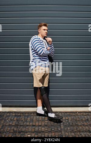 A young man stands thoughtfully with a prosthetic leg, dressed in a striped top and shorts. Stock Photo